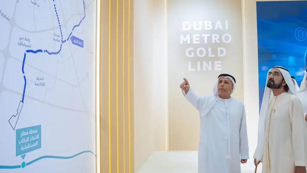 Two men in traditional Emirati dress stand beside a Dubai Metro Gold Line exhibit, one pointing at a large map showing the route.