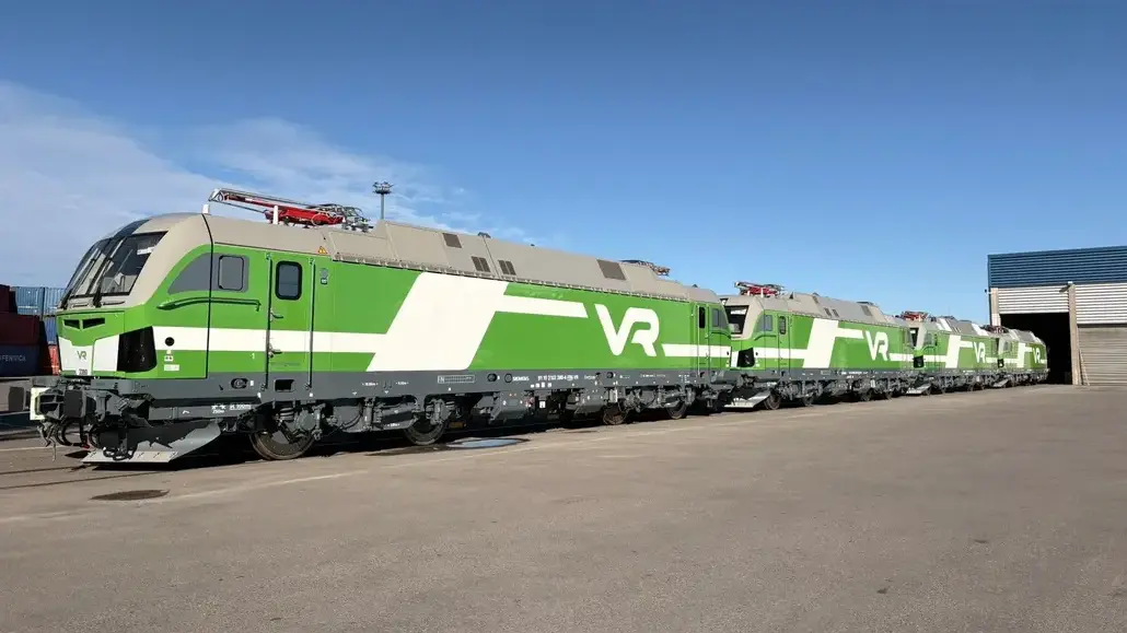 Row of modern VR locomotives in green and white livery parked outside a train maintenance yard under a clear blue sky, extending into the distance