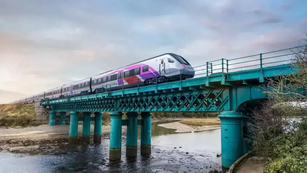 High-speed train crossing a teal metal bridge over a shallow river at sunset.
