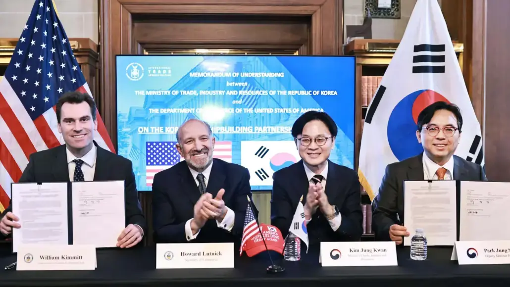 Four men in suits sit at a table signing documents during a memorandum of understanding ceremony, with U.S. and South Korean flags in the background.