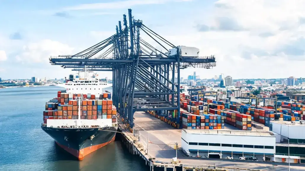 Container ship at a busy port with stacked cargo containers and a large gantry crane overhead.