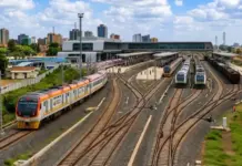 Kenya, France Partner Up for Nairobi Commuter Rail Expansion Wide view of a busy railway station with multiple tracks and trains, including an orange-and-white train on the left and green-and-white trains on the right.