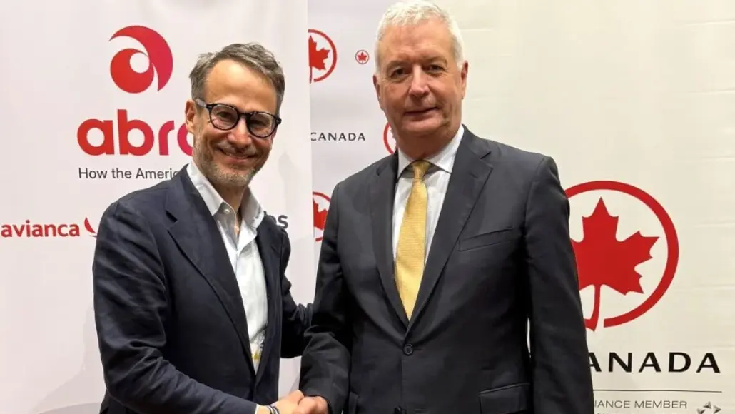 Two men in business suits shake hands at a Canada-themed event backdrop with red logos and banners behind them.