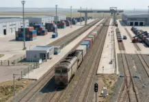 Belarus, Kazakhstan Eye Rail Freight Transport Cooperation Aerial view of a freight rail yard with stacked shipping containers, warehouses, and a diesel locomotive on parallel tracks.