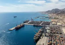 Eritrea, Egypt Discuss Port and Maritime Sector Cooperation Aerial view of a busy container port with colorful stacked containers, cranes, and ships by the blue sea near rugged mountains.