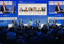 TradeWinds Shipowners Forum Discusses Shipping Challenges Panel discussion at a conference: five panelists seated on a blue stage with a moderator, facing a dark auditorium audience; large screens behind them display slides and headshots.