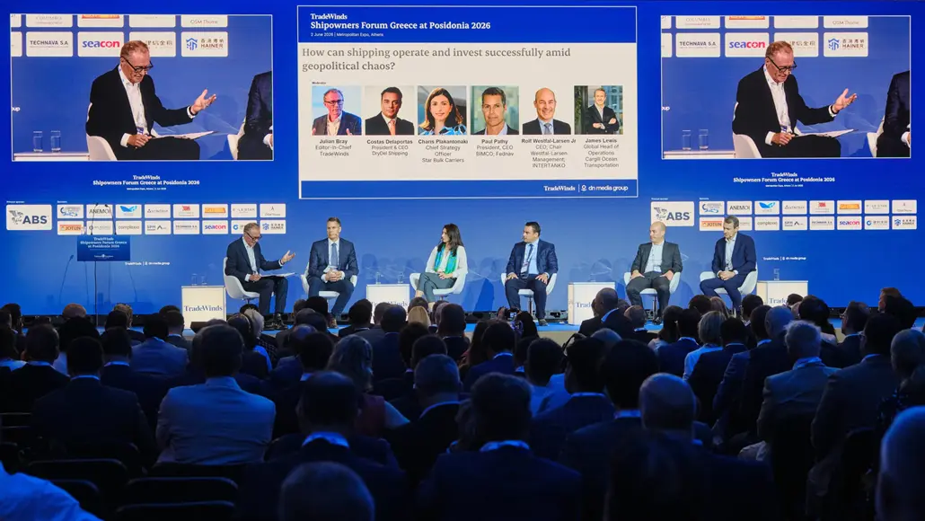 Panel discussion at a conference: five panelists seated on a blue stage with a moderator, facing a dark auditorium audience; large screens behind them display slides and headshots.