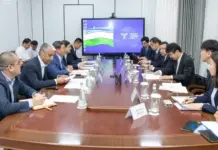 Uzbekistan, JICA Advance Transport Infrastructure Projects Group of professionals in suits seated along a long conference table in a formal boardroom, with a large screen displaying a flag at the far end.