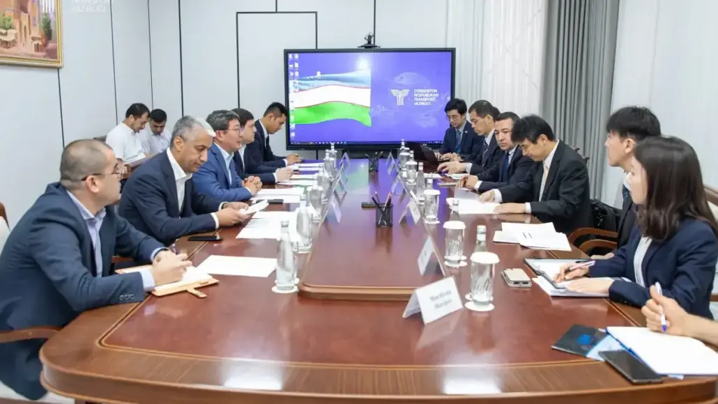 Group of professionals in suits seated along a long conference table in a formal boardroom, with a large screen displaying a flag at the far end.