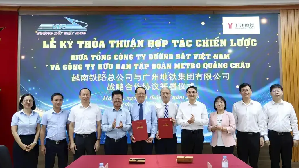Ten professionals in white shirts and blouses pose for a group photo at a signing ceremony, two men in the center hold red folders.