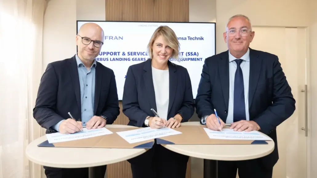 Three business professionals sign documents at a round conference table during a formal signing ceremony, with a screen in the background.