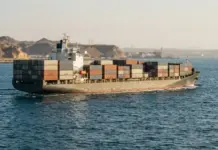 Saudi Arabia’s Red Sea Express Container Line to Boost Trade Container ship sailing through calm blue water with stacked colorful cargo containers and a distant rocky coastline in the background, likely a port route moment.