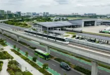 BRICS Adopts Sustainable Transport and Logistics Declaration Aerial view of a modern transit hub with an elevated white tram on tracks above a multi-lane road, green buses on the ground, and a large terminal with aircraft nearby in a cityscape.