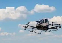 Kazakhstan Debuts Unmanned eVTOL Aircraft Passenger Flight Six-rotor autonomous drone with an enclosed cabin flying in a blue sky with white clouds.
