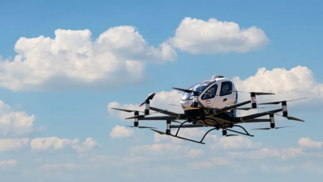 Six-rotor autonomous drone with an enclosed cabin flying in a blue sky with white clouds.