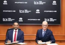 TCS, Porsche Join Hands to Boost AI-Powered Mobility Two businessmen in suits sign documents at a formal signing ceremony, with a backdrop featuring Porsche, TCS, and Tata logos.