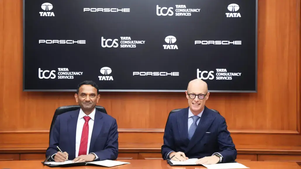Two businessmen in suits sign documents at a formal signing ceremony, with a backdrop featuring Porsche, TCS, and Tata logos.
