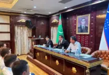 Uzbekistan, Turkmenistan Discuss Boosting Transport and Logistics Ties Three men sit at a gilded conference table addressing an audience in a government-style room with flags and a portrait behind them.