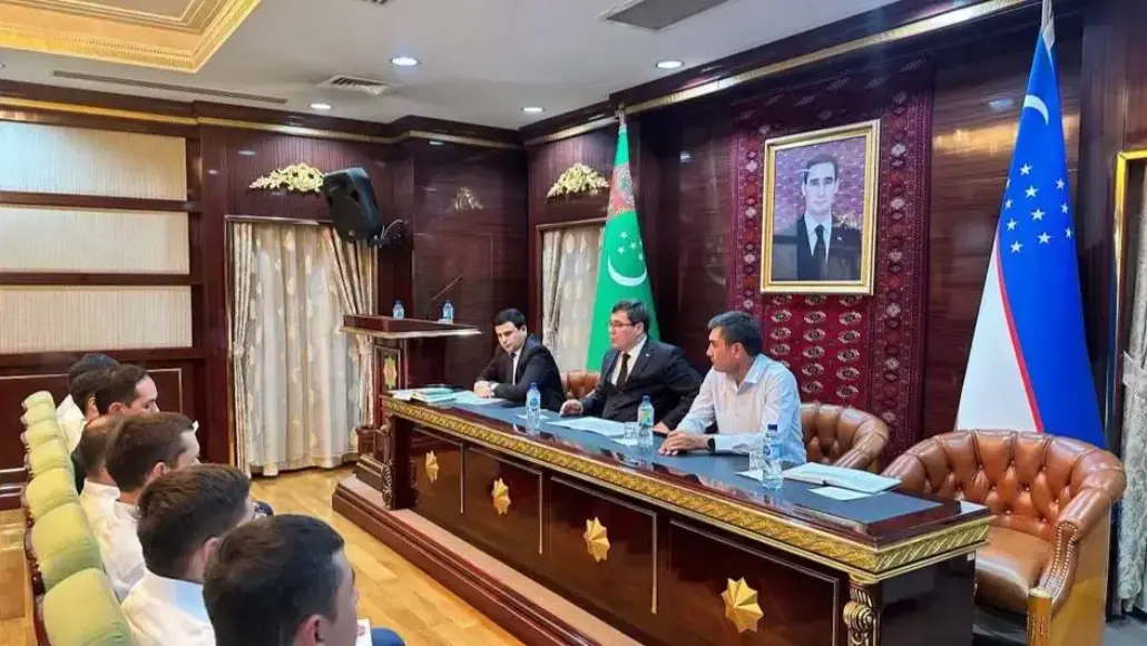 Three men sit at a gilded conference table addressing an audience in a government-style room with flags and a portrait behind them.