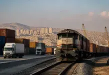 Syria Holds Transport Infrastructure Talks With World Bank Freight train on tracks beside stacked shipping containers at a port, with cranes and a city hillside in the distance.